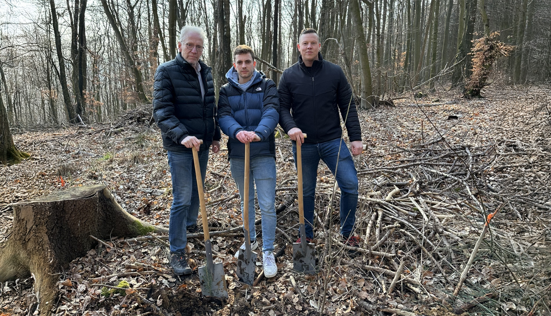 Drei Männer im Wald stehen mit Spaten in der Hand auf Laub, umgeben von Bäumen und abgestorbenem Holz, im Hintergrund befindet sich ein Baumstumpf.