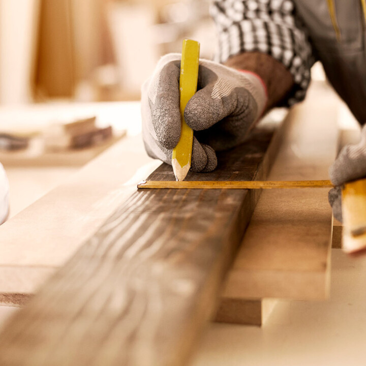 Hand mit grauem Handschuh hält einen gelben Bleistift und markiert ein Maß auf einem dunklen Holzbrett mit einem Zollstock in einer Werkstatt.
